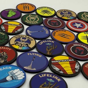 May include: A collection of circular game tokens featuring various colorful graphic designs and text labels including First Strike, Trample, Menace, Vigilance, Hexproof, Flying, Lifelink, Indestructible, Finality, and Reach, with some tokens displaying superhero-themed imagery.