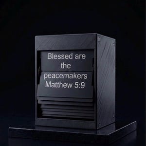 Könnte beinhalten: Eine schwarze, rechteckige Klappzahlenuhr mit strukturierter Oberfläche und weißem Text, der Blessed are the peacemakers Matthew 5:9 lautet. Das Gerät hat eine Reihe horizontaler schwarzer Klappen unter dem Anzeigebereich auf einem festen Sockel.