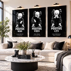 Op de afbeelding: Drie verticale, zwart ingelijste wandkunstprints met een wit stripfiguur met X-vormige ogen. De prints tonen de tekst POSITIVE MIND, POSITIVE VIBES en POSITIVE LIFE, samen met kleinere motiverende zinnen onder elk personage.