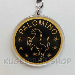 Op de afbeelding: Een ronde acryl sleutelhanger met een zwart midden, een goudkleurige omtrek van een steigerend paard, het woord PALOMINO in gouden letters en kleine gouden sterren. Het ontwerp is omrand door een gouden ring en bevestigd aan een zilveren metalen ring.