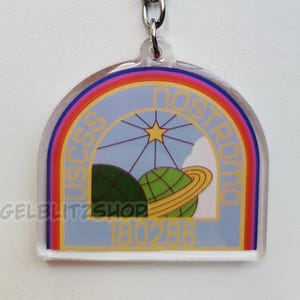 May include: A clear acrylic keychain featuring a semi-circular design with a colorful rainbow border. The graphic depicts a space scene with a yellow star, a green planet with rings, and the text USCSS NOSTROMO 180286. It includes a silver metal ring.
