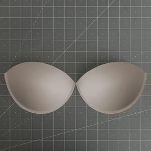 Puede incluir: Un par de copas de sujetador de espuma beige moldeada con una forma suave y redondeada. Las copas están colocadas una al lado de la otra, con un borde superior curvo y un diseño interior cóncavo profundo para la confección de prendas.