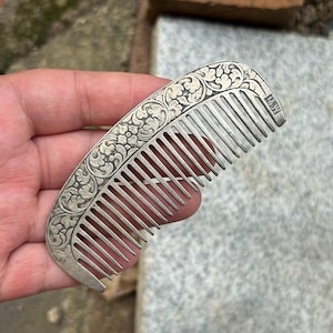 May include: A silver-toned metal hair comb featuring an arched spine decorated with an intricate embossed floral and leaf pattern. The comb has a series of thin, parallel teeth and includes a small rectangular stamp with Chinese characters.