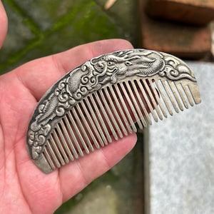 May include: A silver-toned metal hair comb featuring an embossed dragon design along the curved top edge. The comb has a series of thin, parallel teeth and a textured, oxidized finish that highlights the intricate scales and swirling cloud patterns.