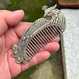 May include: A silver-toned metal hair comb featuring an ornate, raised relief design of a phoenix bird. The handle is shaped like the head and plumage of the bird, with intricate curved lines and spiral details extending down the side.