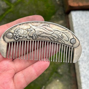 May include: A silver-toned metal hair comb with a curved top edge featuring an embossed relief of a figure in flowing robes. The design includes two small circular accents with characters on the ends and a series of thin, straight teeth.