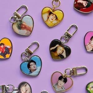 Pode incluir: Uma coleção de porta-chaves em forma de coração com vários retratos fotográficos de pessoas. Cada porta-chaves tem um rebordo metálico em tom dourado ou prateado e um fecho de mosquetão, exibindo fundos de cores variadas como azul, amarelo, vermelho e rosa.