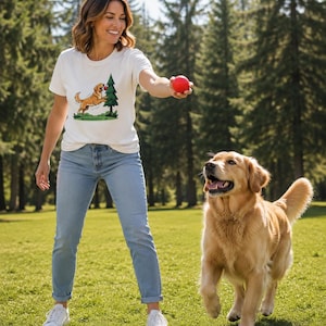 Peut inclure: Un t-shirt blanc à manches courtes orné d'une illustration graphique représentant un golden retriever sautant vers un sapin vert avec une balle rouge. Le haut est associé à un jean en denim bleu clair et des baskets blanches.