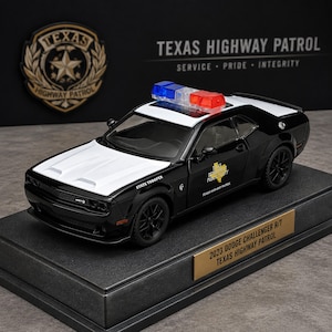 May include: A black and white 2023 Dodge Challenger R/T Texas Highway Patrol model car featuring a red and blue light bar on the roof, black wheels, and the text State Trooper on the front fender, displayed on a base with a gold identification plaque.