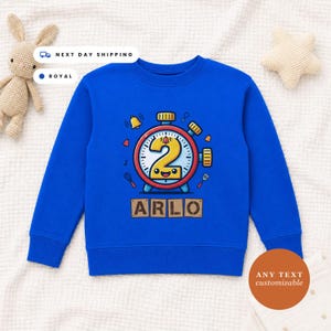 May include: A royal blue long-sleeve sweatshirt featuring a central graphic of a cartoon alarm clock with a large yellow number two on its face. Below the clock, the name ARLO is printed in block letters on a tan background with a distressed texture.
