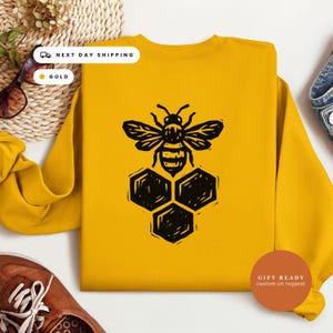 May include: A gold-colored crewneck sweatshirt featuring a black graphic print of a bee positioned above three hexagonal honeycomb cells. The design is centered on the front of the garment, which has long sleeves and a ribbed neckline.