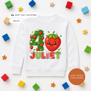 May include: A white long-sleeve sweatshirt featuring a graphic of the number 4, a smiling strawberry character, and the name JULIET in colorful, patterned letters. The number 4 is formed by green strawberry vines with small red berries.