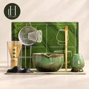 May include: A matcha tea set featuring a green ceramic bowl with a spout, a bamboo whisk in a clear case with Japanese characters, a metal mesh strainer, a bamboo scoop, and a green ceramic whisk holder, all presented in a green box.