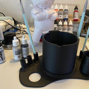 May include: A matte black plastic paint brush and water cup organiser stand featuring a large central cylindrical basin, a smaller side cup, and multiple vertical slots for holding brushes, designed for hobbyist painting and model crafting.