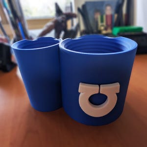 May include: A two-piece blue cylindrical container set with a matte finish. The larger piece features a prominent, raised, off-white omega-shaped emblem on its exterior. Both pieces have a threaded interior design at the top rim for secure closure.