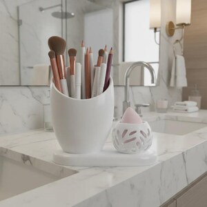 May include: A white ceramic vanity organizer set featuring a tall, curved cup filled with various makeup brushes and pencils, alongside a smaller, spherical holder with a cut-out leaf pattern containing a single pink makeup blending sponge.