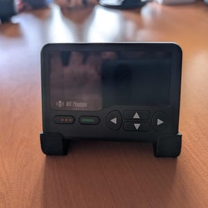 May include: A black WiFi Pineapple network security device featuring a rectangular screen, a directional keypad with four arrow buttons, and two small status indicator lights. The device is mounted in a black plastic stand for upright display.