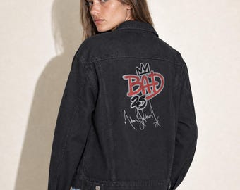May include: A black denim jacket featuring a graphic on the back with the word BAD in red letters, a crown icon above it, the number 25 below, and a signature in white. The jacket has a classic collar, long sleeves, and silver metal buttons.