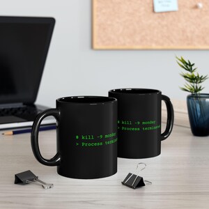 Op de afbeelding: Twee zwarte keramische mokken met groene computerterminaltekst die $ kill -9 monday en > Process terminated leest. De mokken hebben een glanzende afwerking, een gebogen handvat en staan op een lichtgekleurd oppervlak met kantoorbenodigdheden.