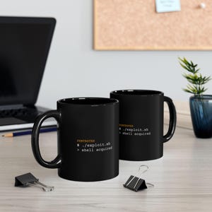 May include: Two black ceramic mugs featuring the orange text PENTESTER followed by the white command line text $ ./exploit.sh and > shell acquired. The mugs have a standard cylindrical shape with a curved handle on the side for easy gripping.