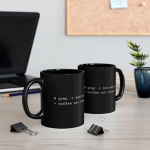 Op de afbeelding: Twee zwarte keramische mokken met witte bedrukte tekst die luidt $ grep -i motivation > coffee not found. De mokken hebben een cilindrische vorm met een gebogen handvat aan de zijkant en tonen dezelfde tekst van de opdrachtregelinterface.