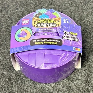 May include: A purple plastic container shaped like a bamboo steamer, featuring a cardboard sleeve labeled The Original Mystery Dumpling Galaxy Edition. The packaging includes text such as Ages 3+, One Set, and The Viral Mystery Dumpling.