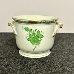 May include: A white porcelain cachepot featuring a green floral motif on the front, gold-trimmed handles, and a green patterned border along the top rim. The vessel has a rounded body, a flared base, and two side handles with gold accents.