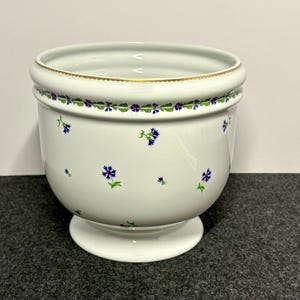 May include: A white porcelain planter featuring a delicate pattern of small blue cornflowers with green stems and leaves. The rim is accented with a thin gold band and a continuous floral border. The vessel has a rounded body and a pedestal base.