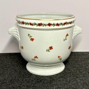 May include: A white ceramic planter with two side handles and a pedestal base. The exterior features a repeating pattern of small orange flowers with green stems and leaves, and a gold-trimmed rim with a floral garland border design.
