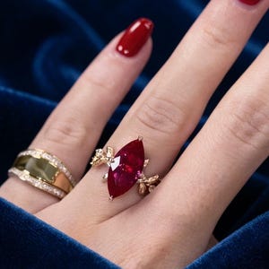 Könnte beinhalten: Ein Ring mit einem tiefroten Edelstein im Marquise-Schliff in einer goldenen Fassung mit filigranen blattartigen Metalldetails an den Seiten, getragen neben einem zweiten Goldring mit breiter Mitteleinlage und kleinen klaren Steinen.