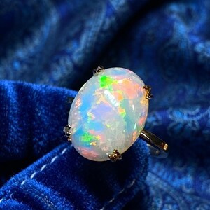 May include: An oval-shaped opal ring featuring a polished, iridescent gemstone with flashes of green, orange, and blue light. The opal is secured by four small metal prongs on a thin, smooth, metallic band that encircles the finger.