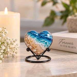 May include: A heart-shaped keepsake urn featuring a resin ocean scene with white crashing waves, sandy beach, and small seashells. Two small starfish are embedded in the sand. The heart rests on a black metal stand with a circular base.