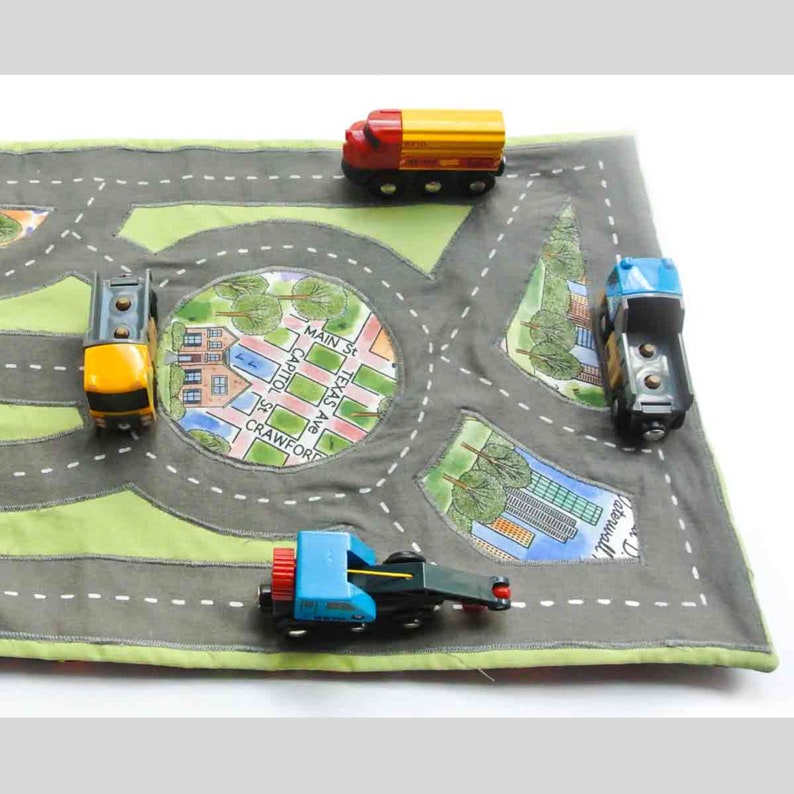 May include: A green and gray play mat with a city map design. The mat features a circular intersection with street names like "Main St" and "Texas Ave". There are four toy trucks on the mat, two blue and two yellow.