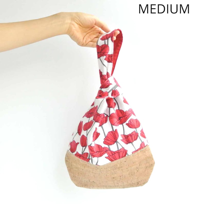 May include: A white fabric bag with a red poppy print and a cork bottom. The bag has a knotted handle and is being held by a hand.