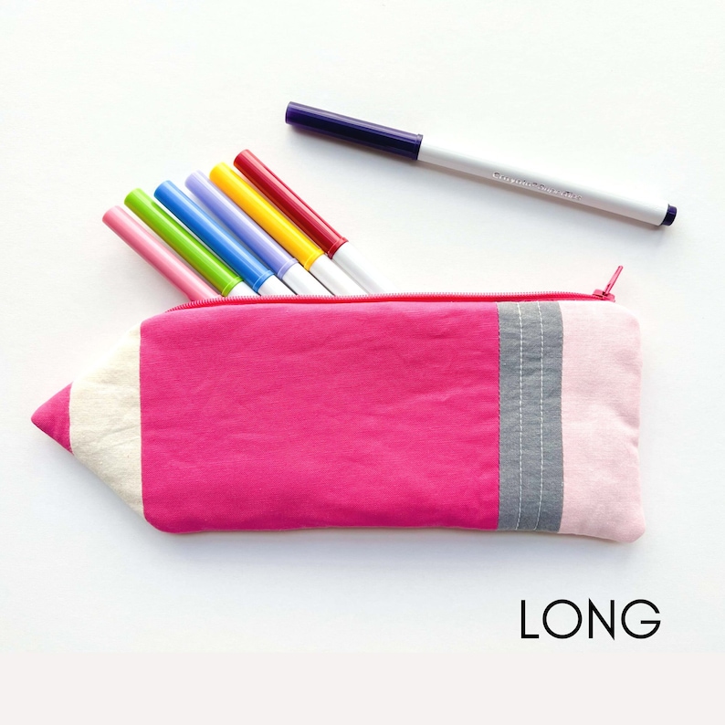 K&ouml;nnte beinhalten: Eine rosa Stiftetui in Form eines Bleistifts mit einer wei&szlig;en Spitze und grauen und rosa Streifen. Das Etui liegt auf einer wei&szlig;en Oberfl&auml;che mit sieben bunten Markern, die aus der Oberseite herausragen.