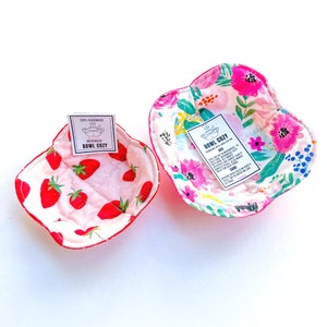May include: Two handmade bowl cozies, one with a strawberry print and the other with a floral print. Both cozies are made with 100% cotton and are microwave safe. The cozies are labeled "Bowl Cozy" and have instructions for use and care.