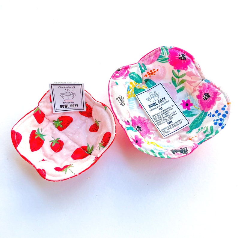 May include: Two handmade bowl cozies, one with a strawberry print and the other with a floral print. Both cozies are made with 100% cotton and are microwave safe. The cozies are labeled "Bowl Cozy" and have instructions for use and care.