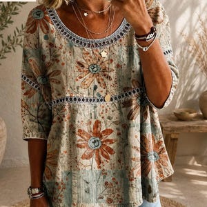 Puede incluir: Una blusa holgada de manga tres cuartos con un estampado floral en tonos apagados, con grandes flores de color naranja amarronado y follaje verde azulado. La prenda presenta un cuello redondo y una banda horizontal con encaje calado decorativo.