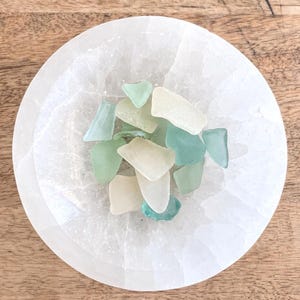 May include: A circular white selenite crystal bowl containing a collection of small, smooth, frosted sea glass pieces in various shades of pale green, aqua, and off-white. The bowl has a translucent, fibrous texture and a polished, rounded edge.