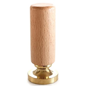 May include: A wax seal stamp featuring a cylindrical light brown wooden handle attached to a circular polished gold-tone metal base. The stamp has a smooth, vertical grain on the wood and a reflective, metallic finish on the bottom sealing plate.