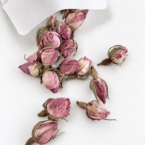 May include: A collection of dried pink rose buds spilling out from a white paper envelope. The rose buds feature varying shades of dusty pink and muted magenta petals, with dried green sepals at the base of each flower head, scattered on a white surface.