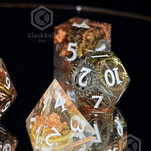 May include: A set of clear resin polyhedral dice featuring embedded metallic gear components and copper-colored foil flakes. Each die has bold white numerals. The logo ClackRoll Dice is visible in the corners of the image, along with a small blue dot.