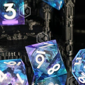 May include: A set of translucent blue and purple polyhedral dice with white numbers. The dice feature internal glitter and are positioned in front of a black, 3D-printed structure with the ClackRoll Dice logo visible on the surface.
