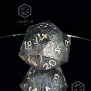 May include: A twenty-sided die made of clear resin with embedded dark glitter and holographic particles. The numbers are engraved in a silver color, with the number 20 clearly visible on the front face. The die reflects on a dark surface.
