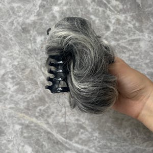 May include: A hair bun extension featuring a mix of grey, silver, and black synthetic hair strands. The hair is styled in a voluminous, wavy texture and is securely attached to a black plastic claw clip with multiple interlocking teeth.