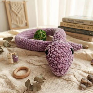 Può includere: Un serpente di peluche realizzato in morbido filato color lavanda testurizzato con occhi in plastica nera liscia. Il serpente è avvolto in una forma circolare, con una testa arrotondata e un corpo lungo e flessibile. La superficie ha un aspetto lavorato a maglia.