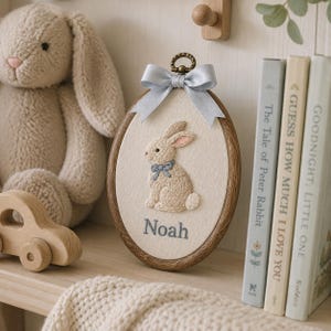 Puede incluir: Un bastidor de bordado ovalado de madera que contiene un panel de tela color crema con un conejo beige bordado que lleva un lazo azul. El nombre Noah est&aacute; bordado en azul debajo del conejo. Un lazo de sat&eacute;n azul claro est&aacute; fijado arriba.