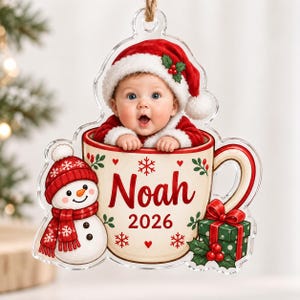 May include: A clear acrylic Christmas ornament featuring a baby wearing a red Santa hat inside a cream-colored mug. The mug displays the name Noah, the year 2026, a snowman, and a green gift box. The ornament has a jute string for hanging.