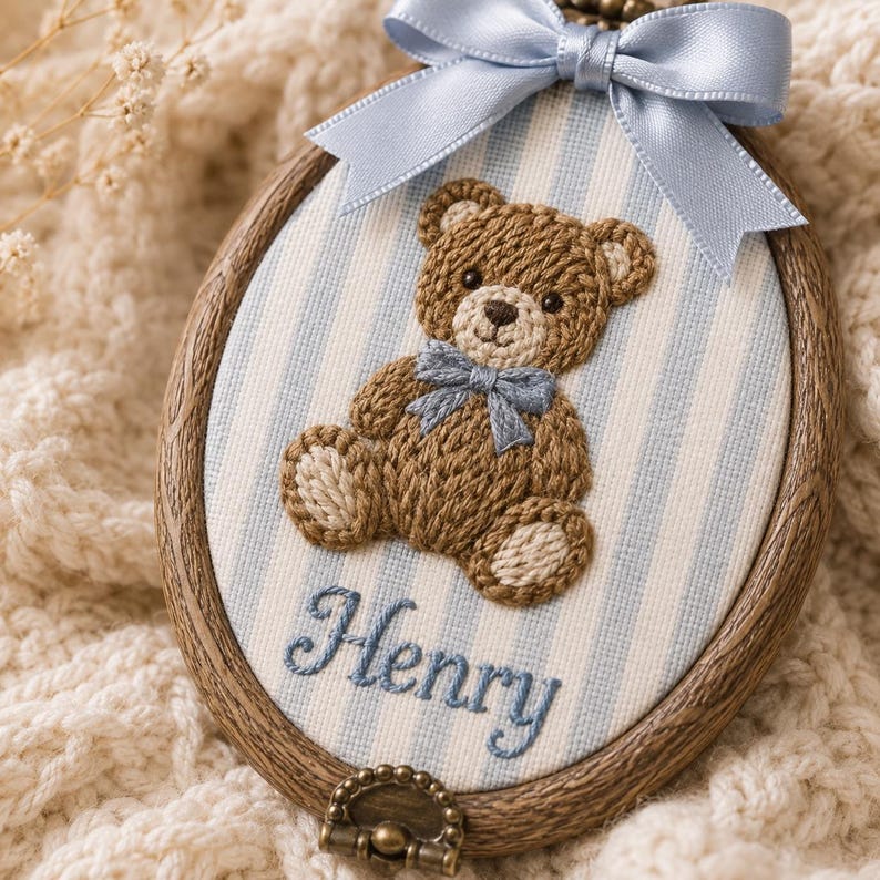 Puede incluir: Un bastidor de bordado ovalado de madera que contiene un panel de tela con rayas verticales azul claro y blancas. Un oso de peluche bordado en marr&oacute;n con pajarita azul se sit&uacute;a sobre el nombre Henry bordado. Un lazo de sat&eacute;n azul est&aacute; unido.