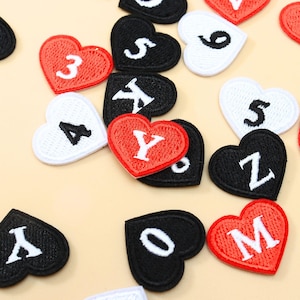 Puede incluir: Una colección de parches bordados en forma de corazón en rojo, negro y blanco. Cada parche presenta una letra o número bordado en un color contrastante. Los parches están dispersos, mostrando varios caracteres, incluidos 3, 4, 5, 6, Y, Z, M y O.